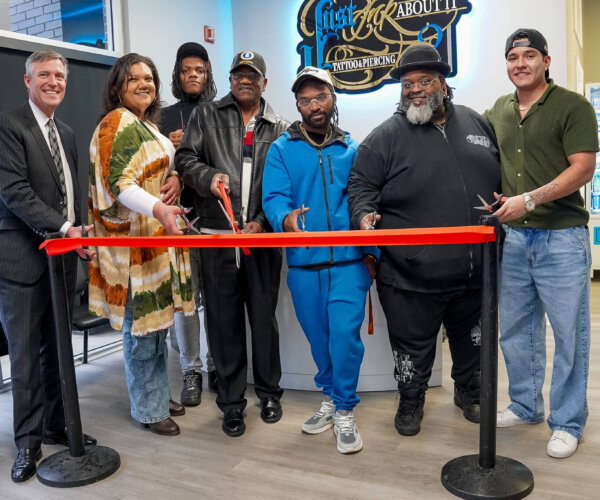 Picture of people standing behind a red ribbon, about to cut it for the grand opening of Just Ink About It Tattoo Studio