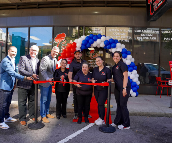 Picture of people standing behind a red ribbon, about to cut it for the grand opening of Cuban Corner